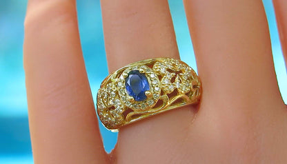 A gold ring with an oval blue sapphire center stone and small clear diamonds set in a flower pattern, shown worn on a finger against a bright blue background.