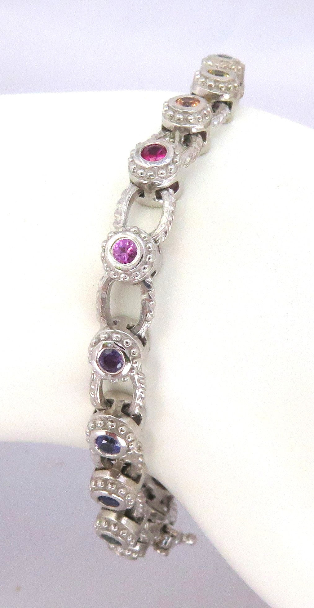 A white gold link bracelet displayed on a white mannequin arm, featuring alternating oval links with round pink, purple, and blue sapphires