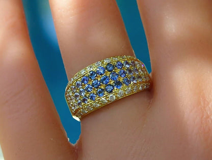 A close-up of a yellow gold  pavé ring with blue sapphires and clear accent diamonds, worn on a finger against a bright blue background.