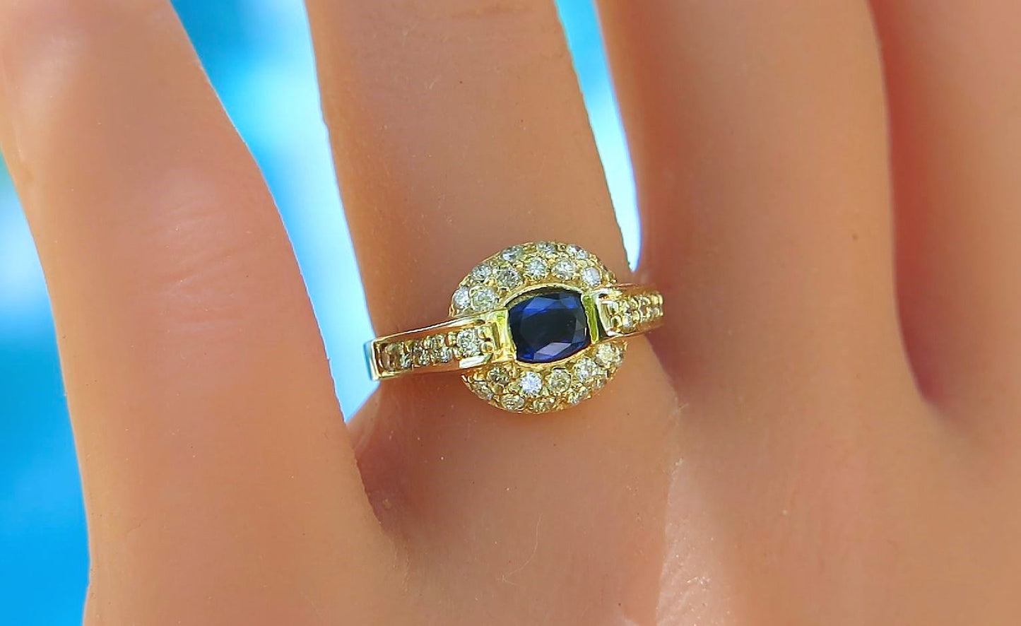 A yellow gold ring with a central blue sapphire oval gemstone surrounded by a halo of small clear stones, shown worn on a finger.