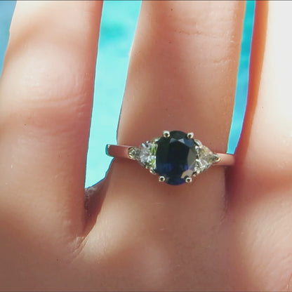 3 stone ring with oval  blue sapphire center and trillion white sapphire side stones in simple white gold mounting on the hand with a turquoise background