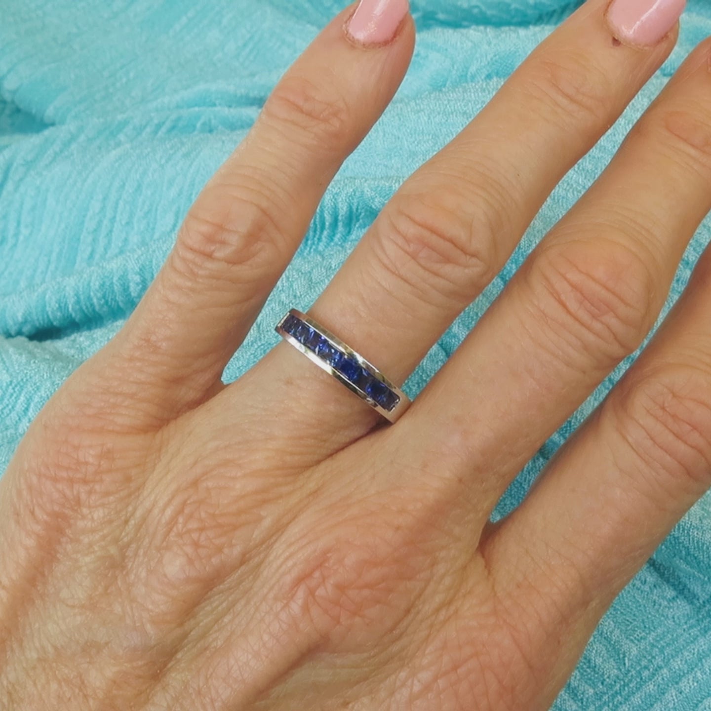 channel set princess cut blue sapphire band in 14k white gold shown on the hand with a blue background