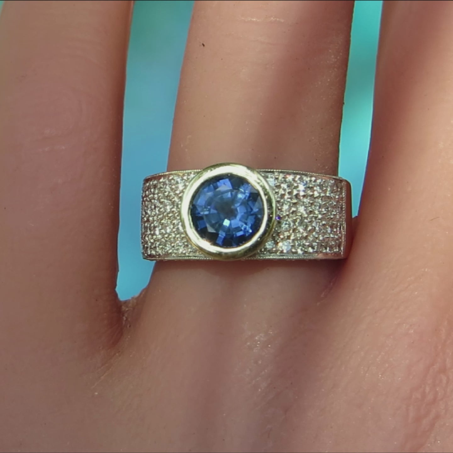 wide band ring with round bezel set blue sapphire and pave diamonds in white and yellow metal on ladies' hand