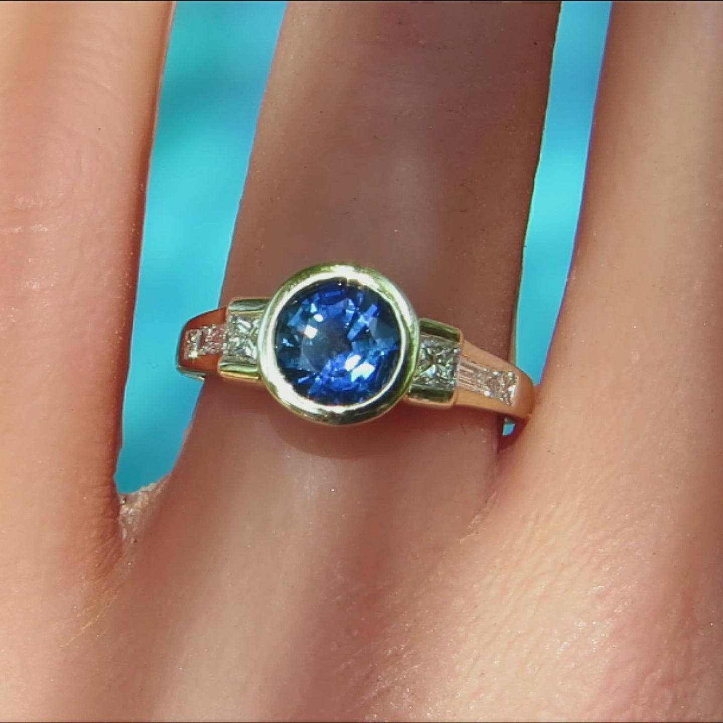 Bezel set blue sapphire ring with princess and baguettte clear diamonds channel set in the band in white and yellow metal on a ladies finger