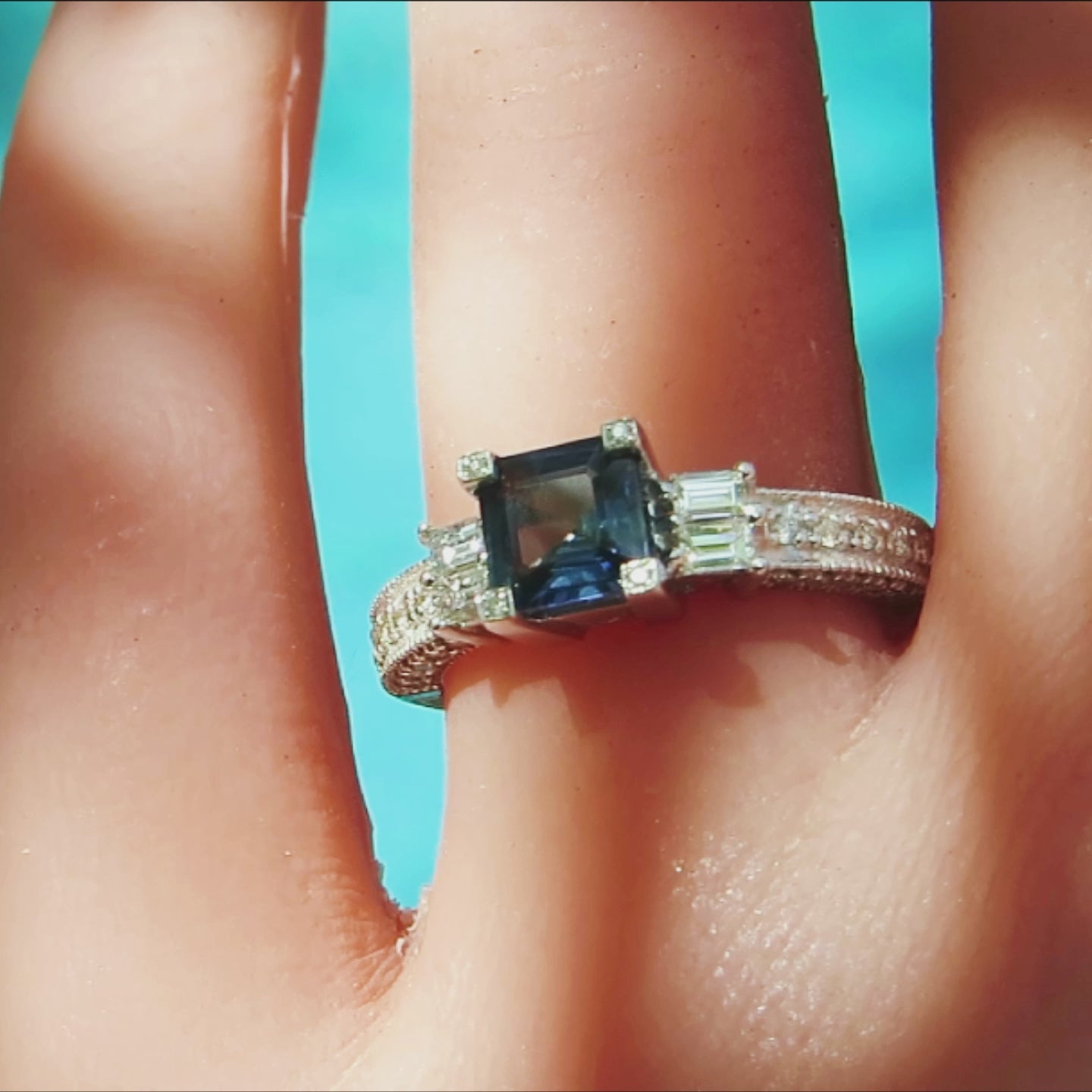 square cut blue sapphire with diamond accented prongs flanked by pairs of baguette diamonds in white gold on the finger