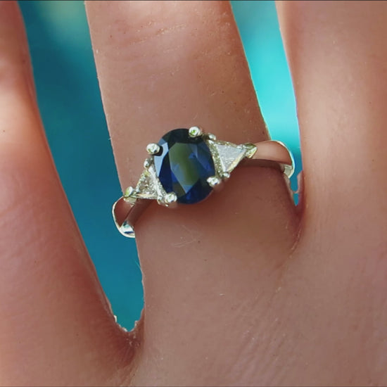 White gold ring with three stone setting featuring oval blue sapphire flanked by trillion white diamonds on the finger