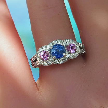hand view of round blue sapphire and pink sapphire ring encrusted with fine diamonds in a white metal 14k on a ladies hand with blue background