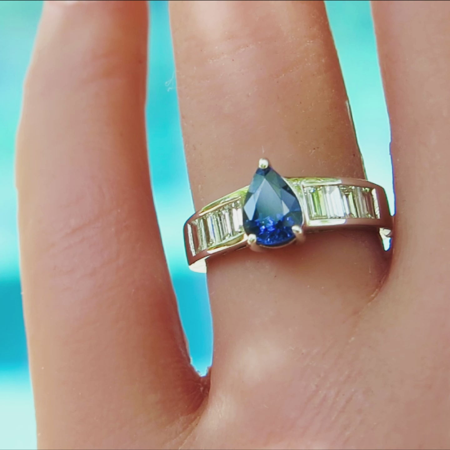 sleek modern pear cut  blue sapphire prong set flanked by channel set baguette diamonds in 14k white gold on ladies' finger with bright blue background