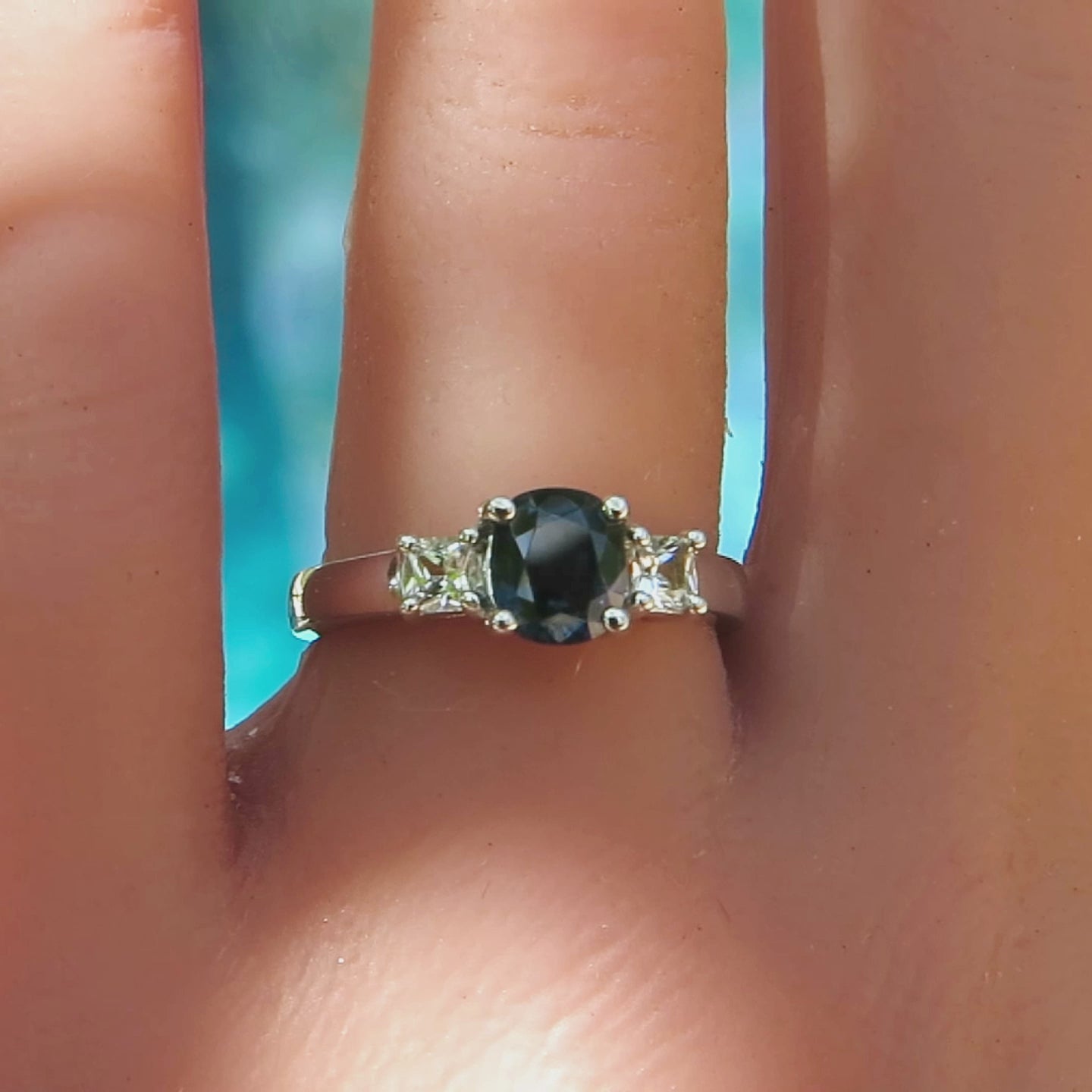 3 stone ring with cushion cut blue sapphire and princess cut white sapphires in white metal ring on the finger with blue background