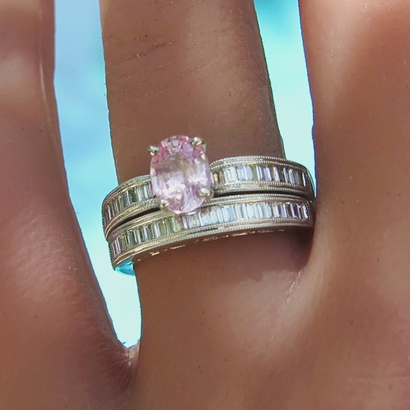 pastel pink oval sapphire with channel set baguette diamonds engagement ring and matching band in white metal on the hand