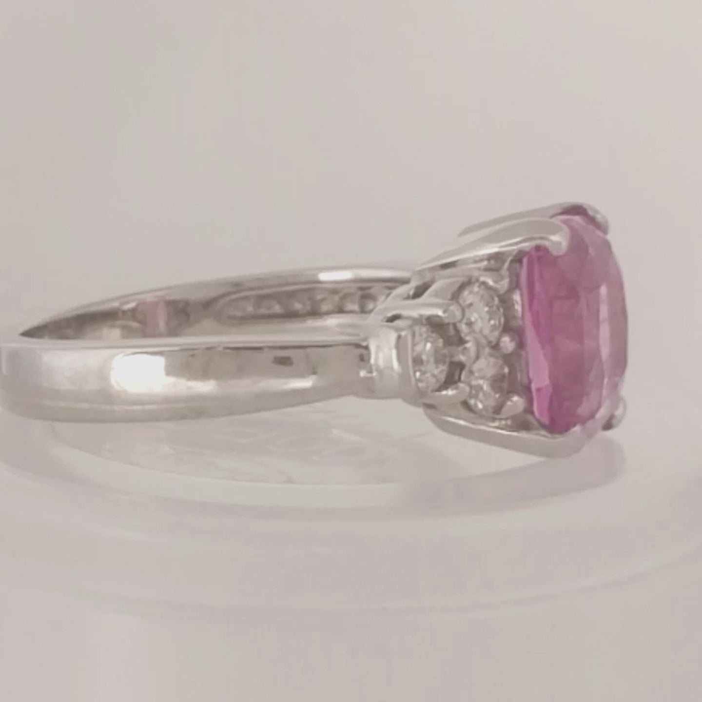 14k white gold ring with cushion cut pink sapphire at the center and 3 round diamonds on either side on a white turntable