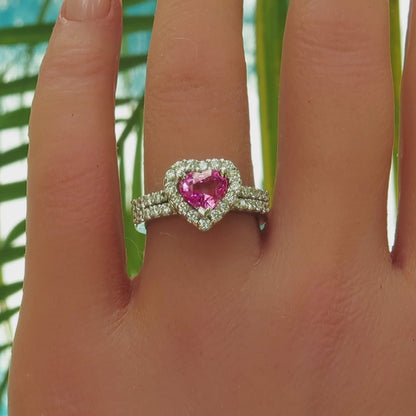 Heart shape bright pink sapphire diamond halo engagement ring and wedding band in white metal platinum on the hand with palm leaves in the background