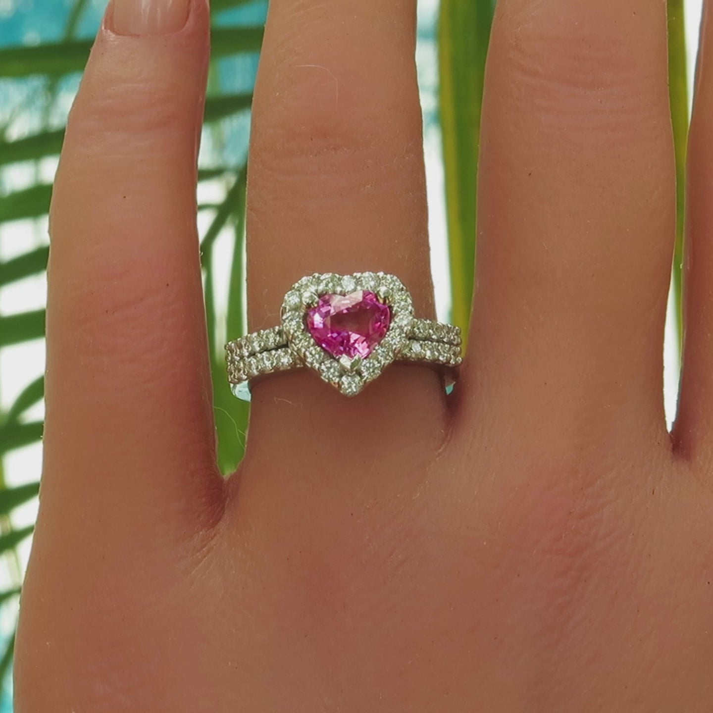 Heart shape bright pink sapphire diamond halo engagement ring and wedding band in white metal platinum on the hand with palm leaves in the background