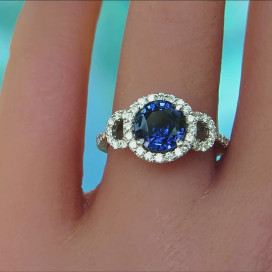 sapphire ring with diamond halo design setting in white metal on a ladies finger with a blue background