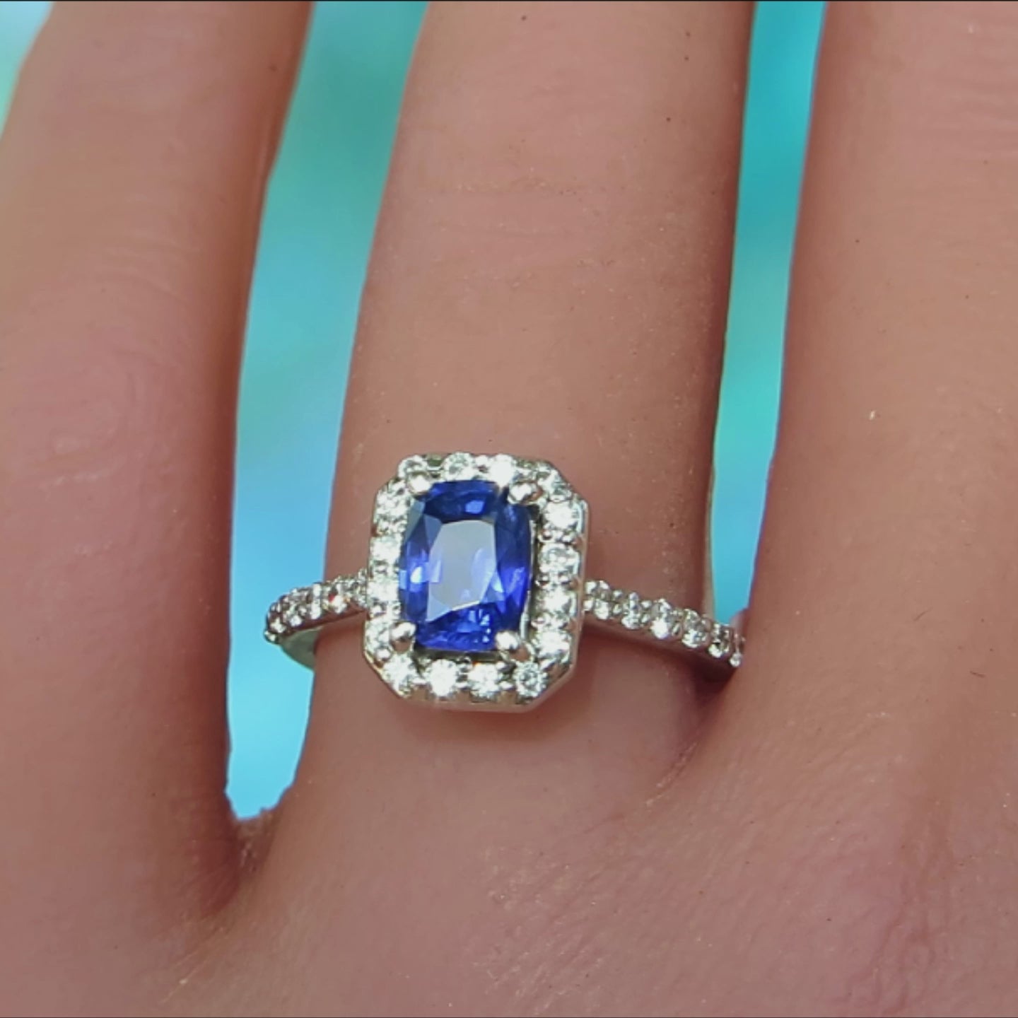 cushion cut blue sapphire surrounded by clear diamonds and diamonds in the band in a white metal on the finger