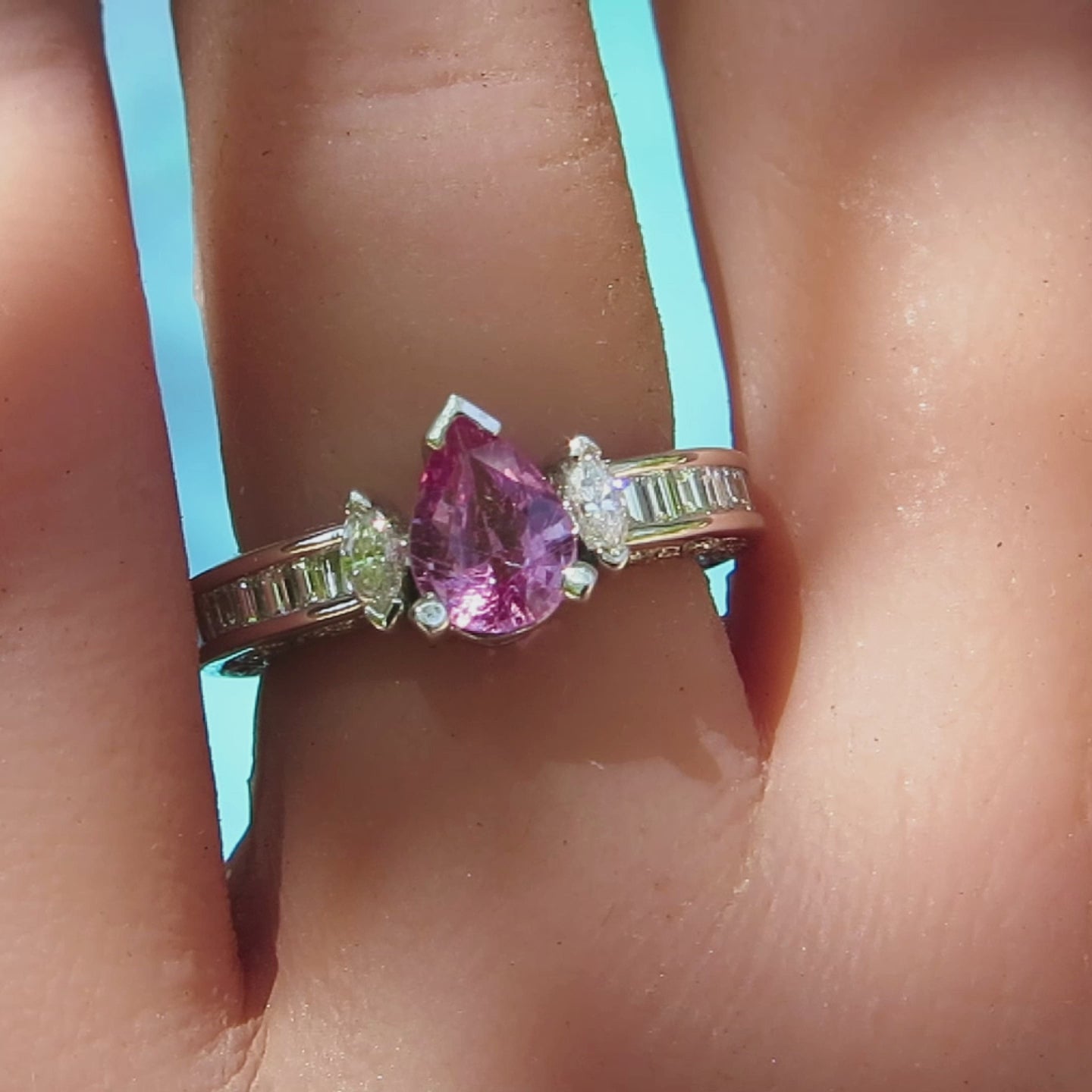 pear cut pink sapphire ring with marquise side diamonds and baguette diamonds channel set in an eternity style ring in white gold shown on the hand