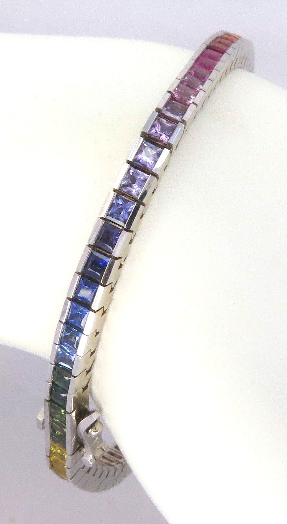 A white gold tennis bracelet featuring a continuous row of princess-cut rainbow-colored gemstones, displayed on a white background.
