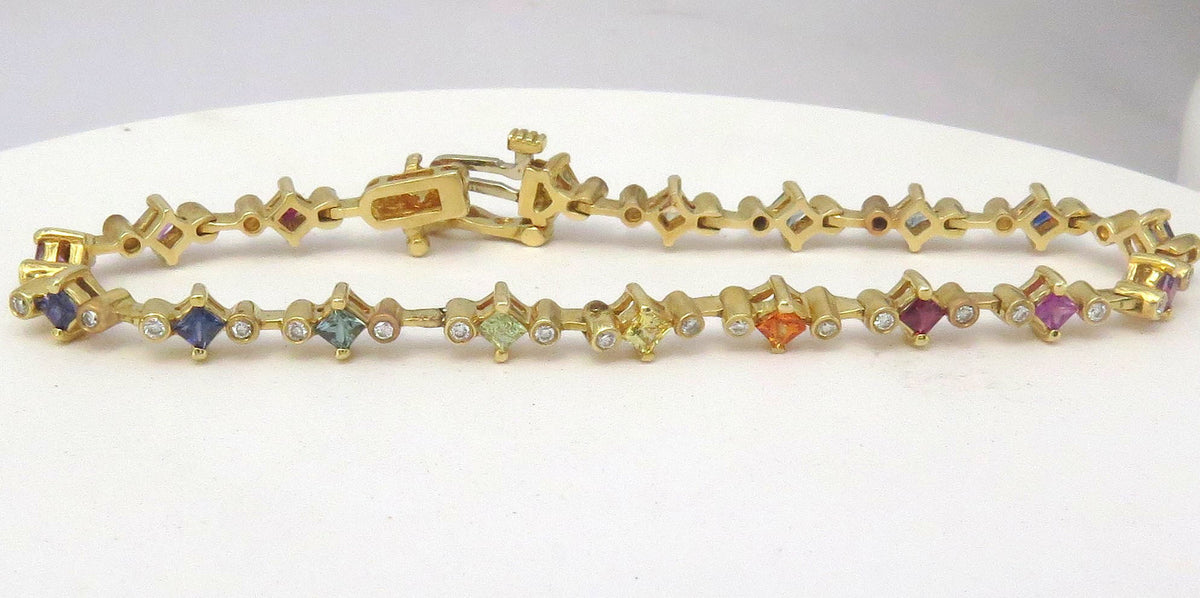 A yellow gold tennis bracelet featuring small diamond-shaped links set with various colored sapphires and tube set diamonds, displayed on a white surface.