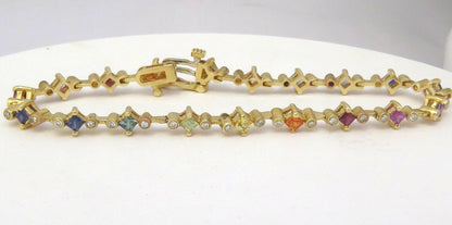 A yellow gold tennis bracelet featuring small diamond-shaped links set with various colored sapphires and tube set diamonds, displayed on a white surface.