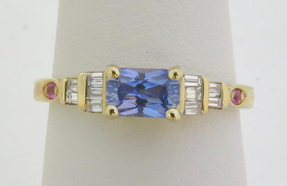 A yellow gold ring featuring a radiant cut blue sapphire center stone, flanked by baguette-cut clear diamonds and small round pink gemstones on a white ring stand.