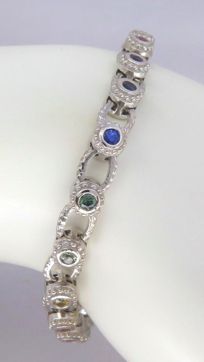 A white gold link bracelet displayed on a white mannequin arm, featuring oval links with round sapphires in blue, green, and yellow.