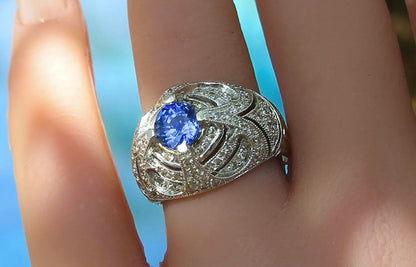 A bold white gold filigree ring with a round blue sapphire gemstone, worn on a finger against a bright blue background.