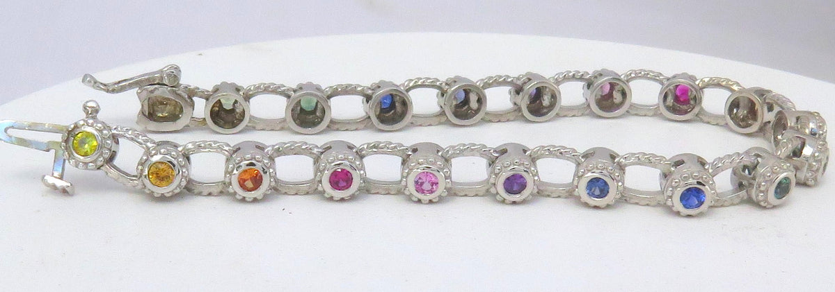A white gold tennis bracelet with multiple round-cut rainbow sapphires in various colors including pink, blue, yellow, and green, displayed on a white background.