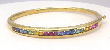 A gold hinged bangle bracelet featuring a row of small, multicolored sapphires set along the front, displayed on a plain white background.