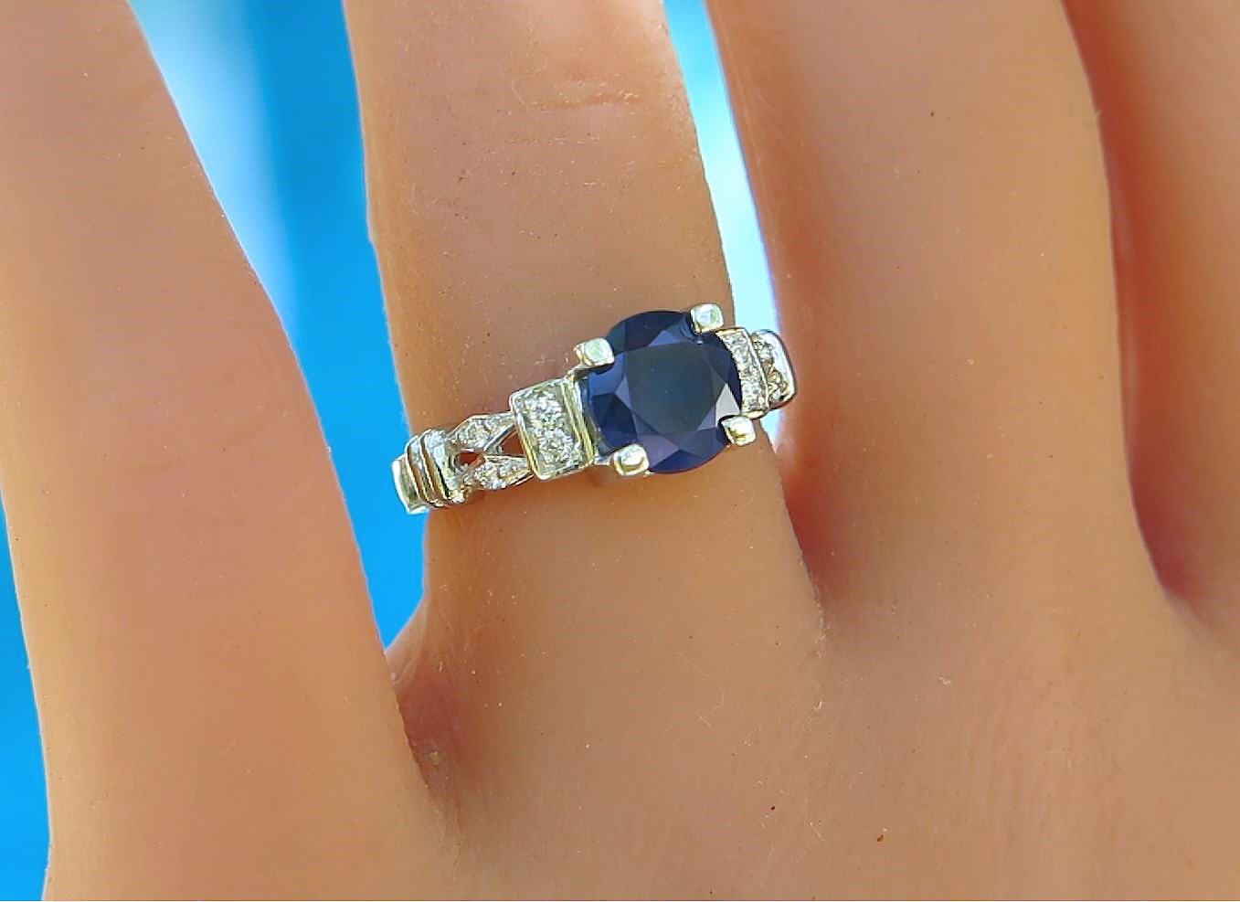 A 14k white gold ring with a round dark blue sapphire center stone and small clear diamonds, shown worn on a finger against a bright blue background.