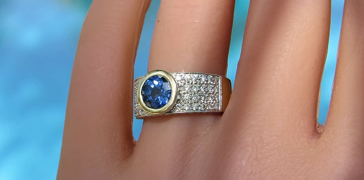 A close-up of a gold ring with a round blue gemstone and a pavé-set diamond band, worn on a finger against a blurred blue background.