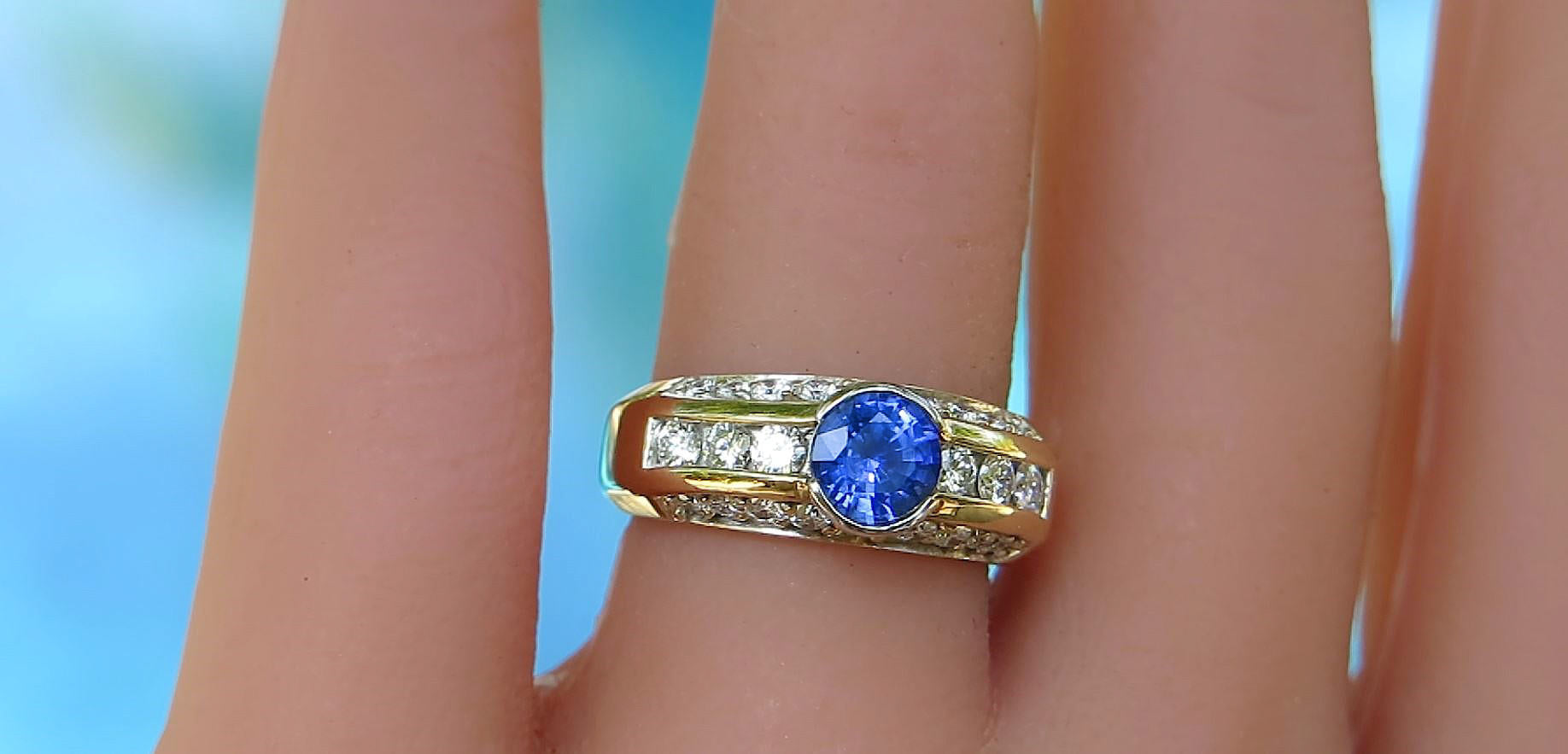 A close-up of a gold ring with a round blue gemstone and channel-set clear stones, worn on a finger against a soft blue background.