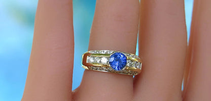 A close-up of a gold ring with a round blue gemstone and channel-set clear stones, worn on a finger against a soft blue background.