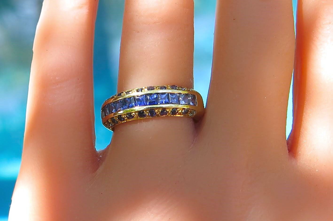 A yellow gold ring featuring a channel-set row of round blue sapphires and small round diamonds, shown worn on a finger against a blurred blue background.