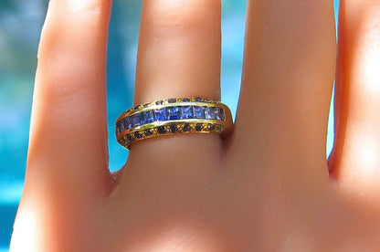 A yellow gold ring featuring a channel-set row of round blue sapphires and small round diamonds, shown worn on a finger against a blurred blue background.
