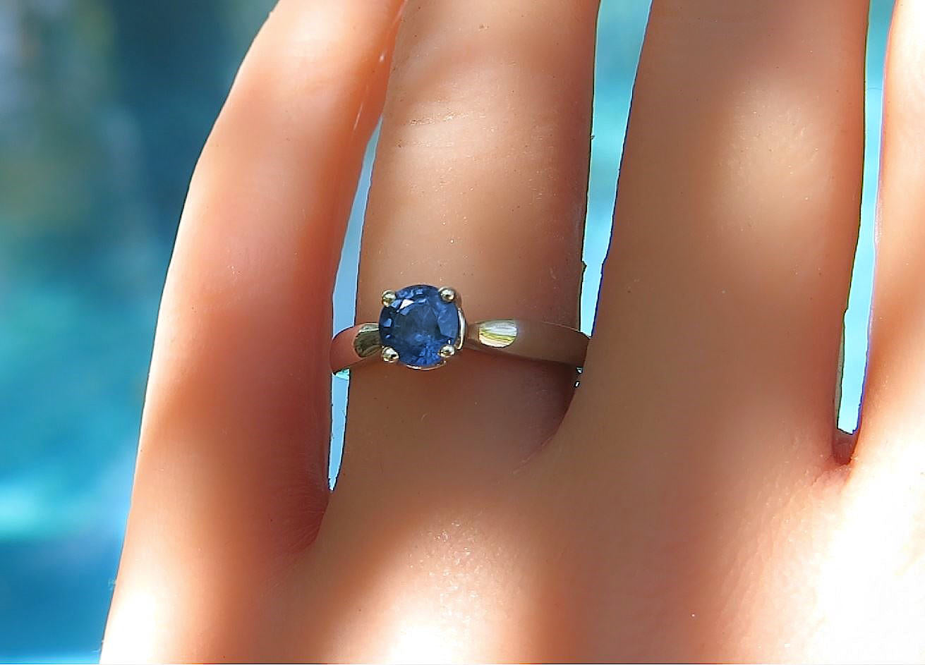 A white gold ring with a round blue gemstone set on a finger, photographed close-up against a blurred blue background.