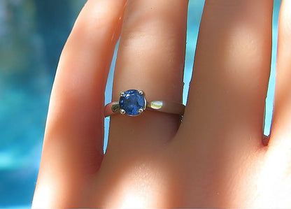 A white gold ring with a round blue gemstone set on a finger, photographed close-up against a blurred blue background.