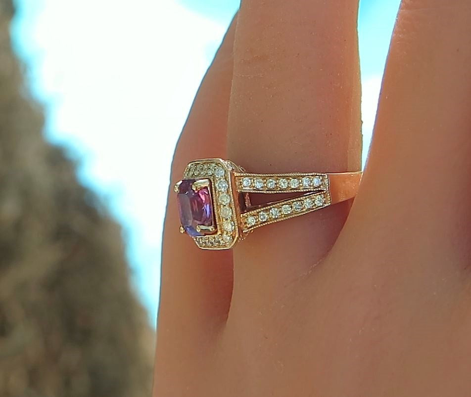 A close-up shot of a rose gold ring with a square purple magenta sapphire and small clear accent diamonds, worn on a finger against a blurred outdoor background.