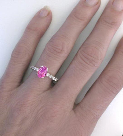 A silver-toned ring with a prominent oval pink gemstone and small clear accent stones, shown worn on a person's finger against a plain light background.