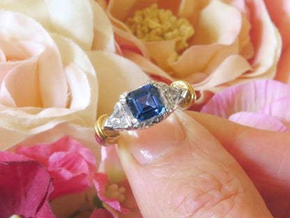 A close-up of a hand wearing a ring with a blue gemstone and diamond accents on a gold band, set against a background of soft pink and cream roses.