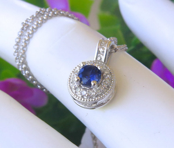 Round natural blue sapphire and diamond halo pendant in white gold on a finger with floral background