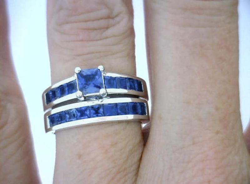 A close-up of a hand wearing a silver ring set with deep blue sapphires, featuring a square-cut central sapphire and matching channel bands.