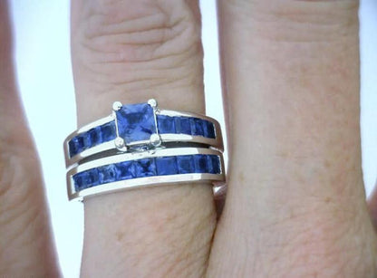 A close-up of a hand wearing a silver ring set with deep blue sapphires, featuring a square-cut central sapphire and matching channel bands.