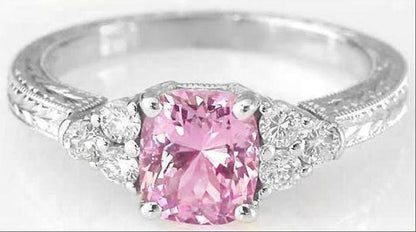 A silver-colored ring featuring a cushion-cut pink gemstone in a prong setting, accented by small clear stones on the shoulders, shown on a plain white background.