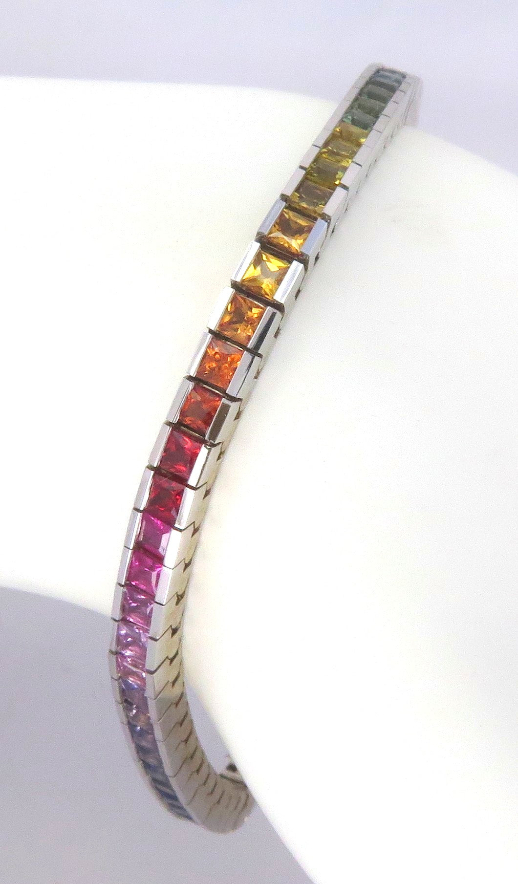 A white gold tennis bracelet with a channel set rainbow sapphires, shown in a close-up studio shot against a white background.