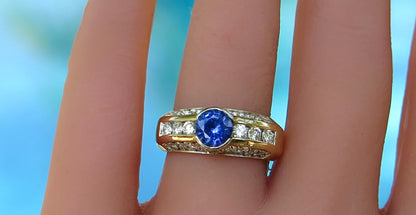 A yellow gold ring with a round blue sapphire center stone and small clear diamond accent stones, shown worn on a finger against a blurred blue background.