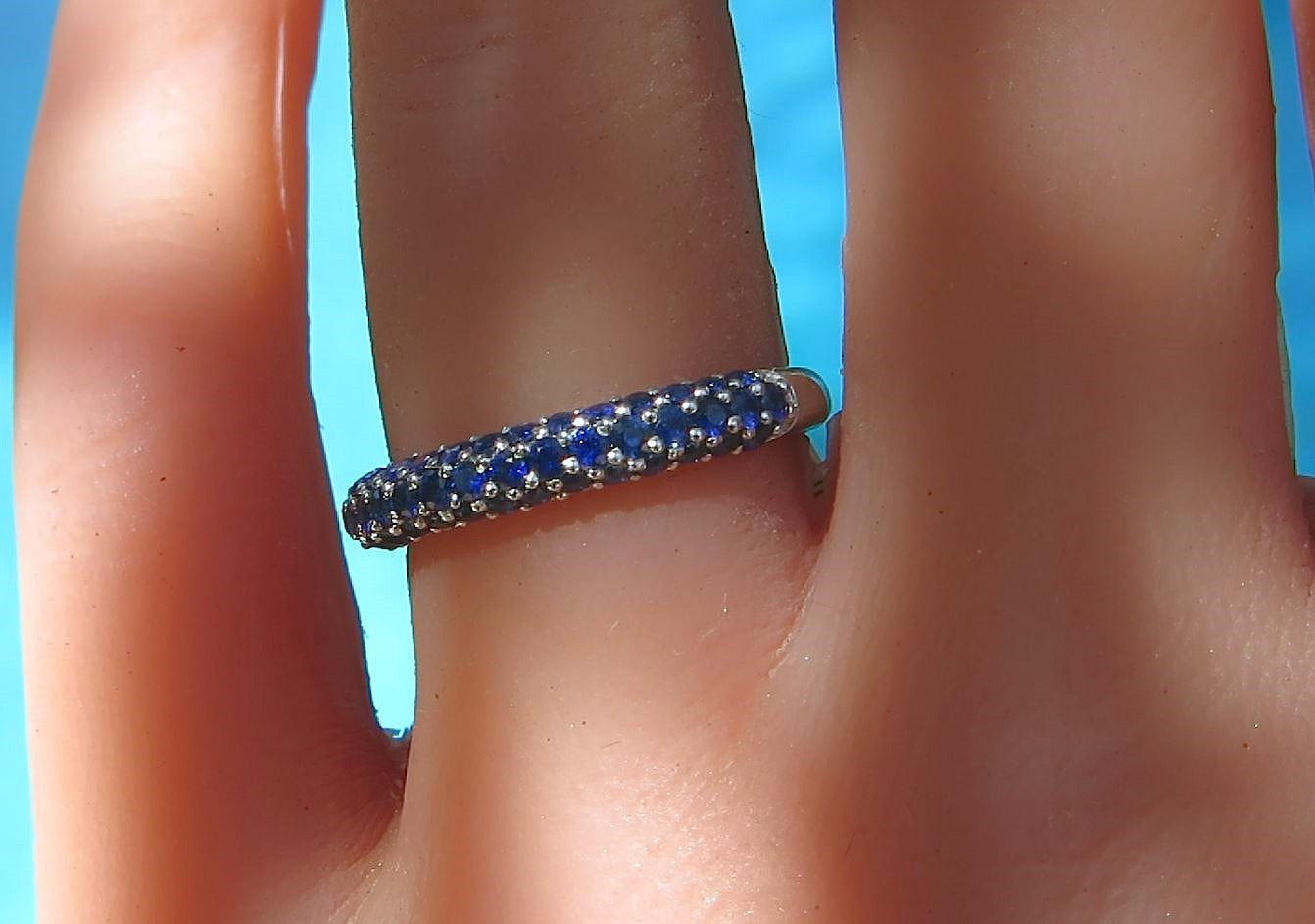 A close-up of a ring worn on a finger, featuring a white gold band set with deep blue round sapphire against a bright blue background.