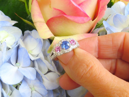 A hand holding a white metal ring with a central blue sapphire and pink sapphires, surrounded by light blue hydrangeas and a pink rose.