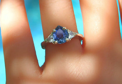A close-up of a ring worn on a finger, featuring a central blue pear-cut gemstone with triangular clear side white sapphires and a white-gold-toned band, set against a bright blue background.