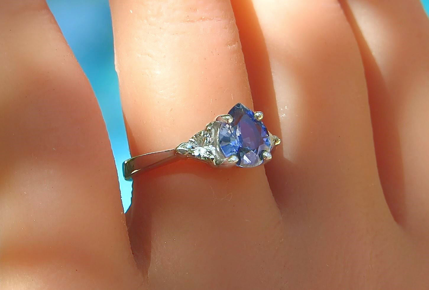 A close-up of a white gold ring with a pear cut blue gemstone and small clear accent white sapphires, worn on a finger.