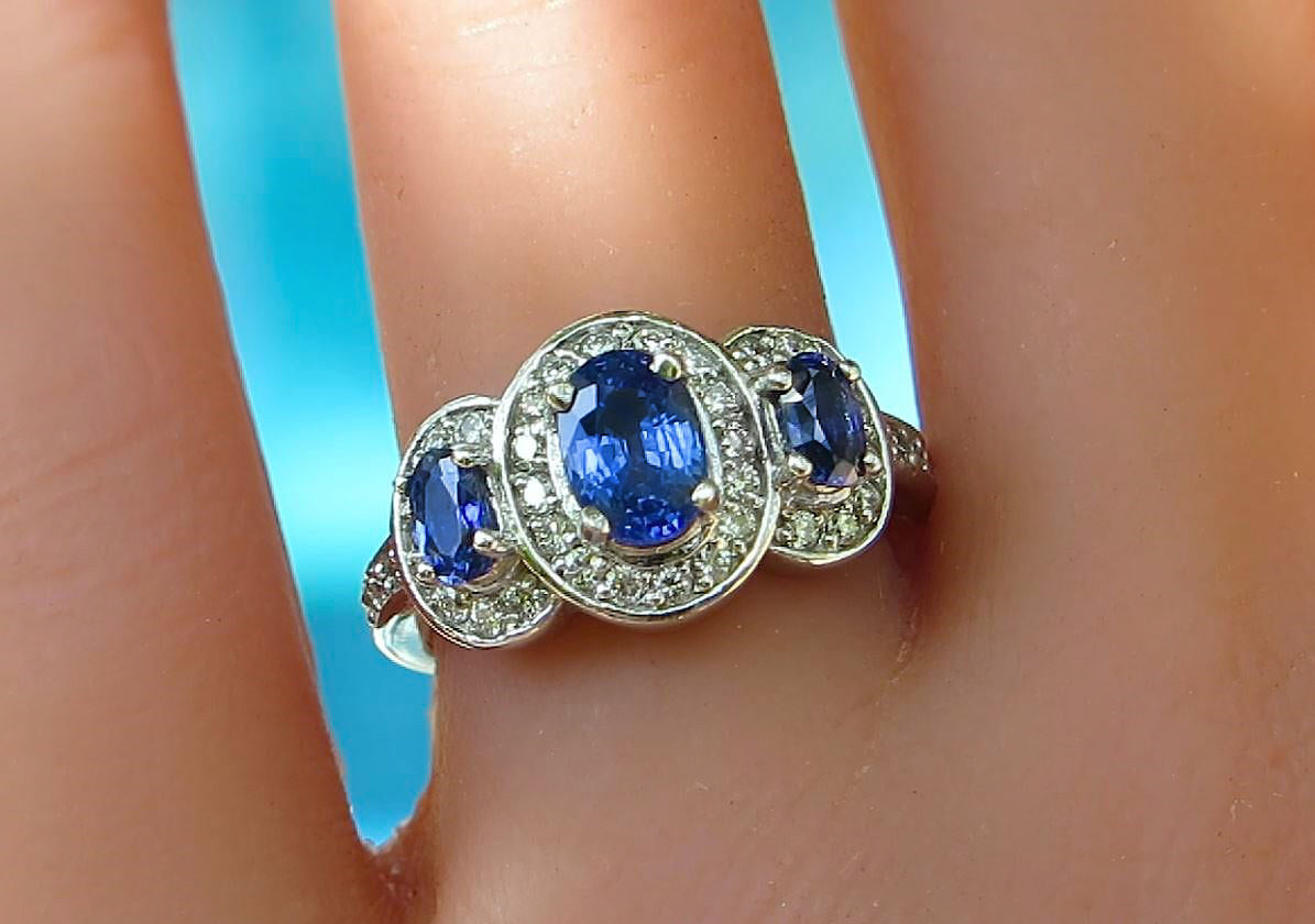 A white gold ring with three oval blue gemstones and small clear accent diamonds , shown worn on a finger against a bright blue background.